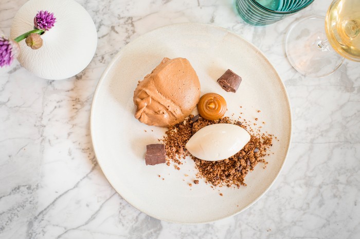 A white plate against a marble background. On the plate is a pale chocolate mousse with a scoop of ice cream and crumble biscuit base