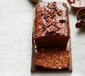 A dark brown Date and pecan loaf cake with chocolate bitters on a small board with a white background