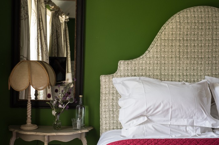 A bedroom with pea green walls, a cream fabric head board and a side table with flowers on