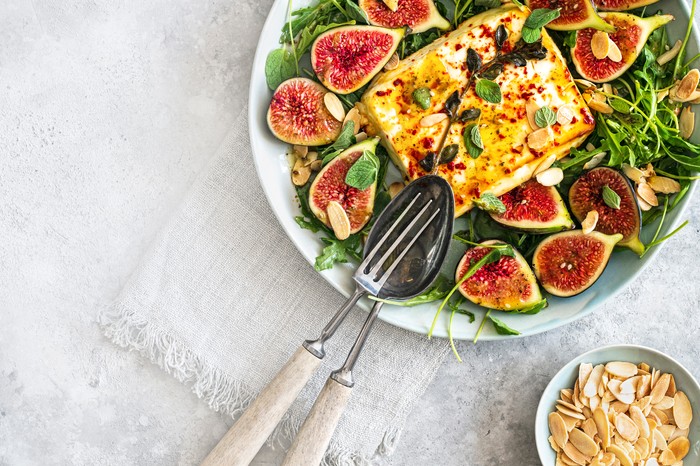 A large white serving plate filled with green leaves and topped with halved figs and a block of baked feta, on a light stone background