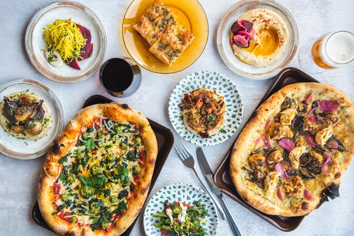 Two oval shaped pizzas topped with broccoli and cauliflower, next to six small plates such as hummus, focaccia, pickles and broccoli