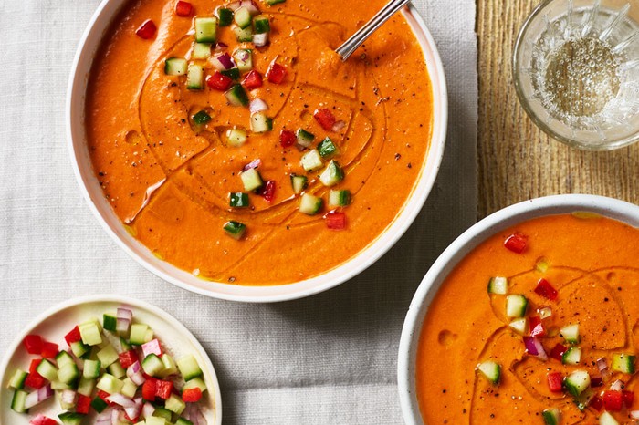 Two bowls of red gazpacho with a side dish of chopped cucumber and tomato