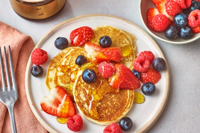 A white plate with three pancakes topped with sliced strawberries and blueberries