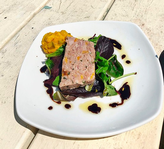 A white plate of ham hock terrine with salad leaves on a wooden garden table in the sun