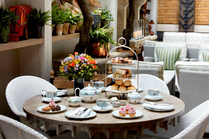 A table laid out for afternoon tea at Ham Yard Hotel including a three tiered cake stand, a blue and white tea pot and three blue and white tea cups and saucers, with flowers in pots in the background