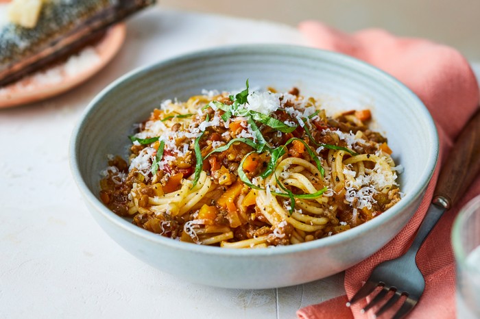 A white bowl of spaghetti bolognese with parmesan and a grater in the background