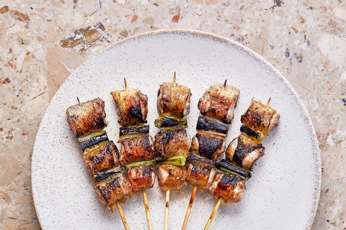 A white mottled plate on a stone background with five bamboo skewers fanned out, threaded with charred chicken