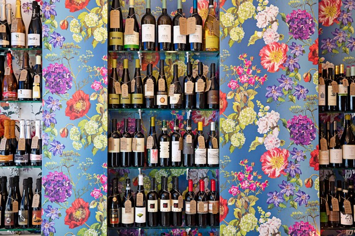 A colourful room has floral-patterned wallpaper. Between the wallpaper is a section with bottles of wine