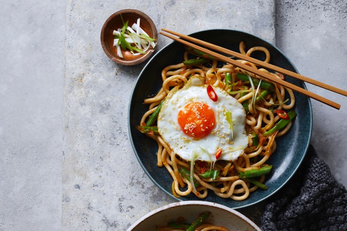 Kimchi Noodles A navy-blue bowl of thick noodles with vegetables and a fired egg on top, and chopsticks resting on the bowl