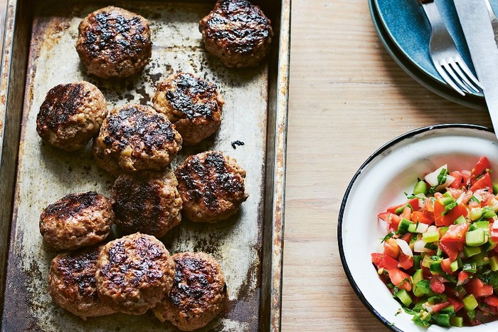 A silver oven tray of round beef and lamb patties next to a chopped salad