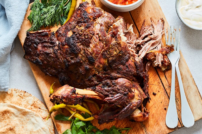 A lamb joint on a wooden board with a pot of red chunky sauce, a couple of forks and some flatbread