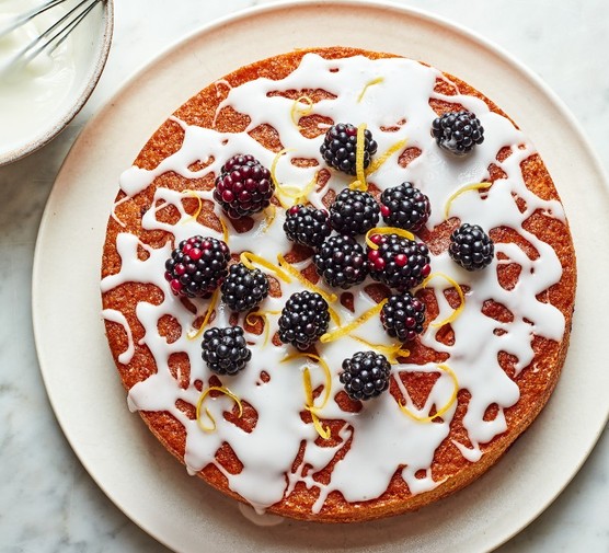 A sponge cake topped with splattered icing sugar and a scattering of blackberries on a white plate