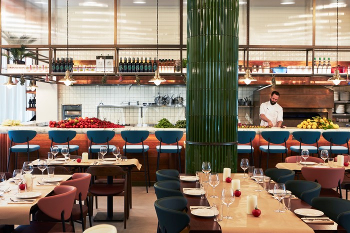 A restaurant with tables laid, an emerald pillar and an open kitchen with a chef chopping