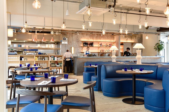 A restaurant room with a large concrete bar at the back with wooden tables and blue banquette seating