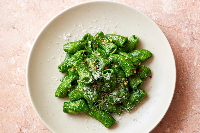 A white plate topped with rigatoni pasta coated in a vibrant green sauce and sprinkled with chilli flakes