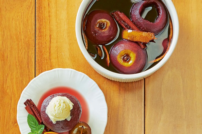 Poached apple on a plate topped with ice cream next to cinnamon sticks and mint. Next to a bowl of more poached apples with orange zest