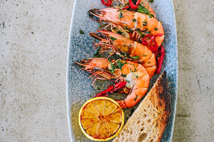 A rectangular plate topped with four prawns with their shells on and half a lemon