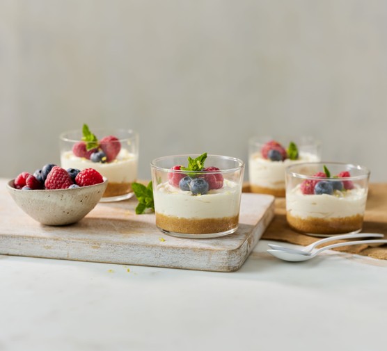 Four pots of mini cheesecakes topped with summer berries next to a small bowl of more berries