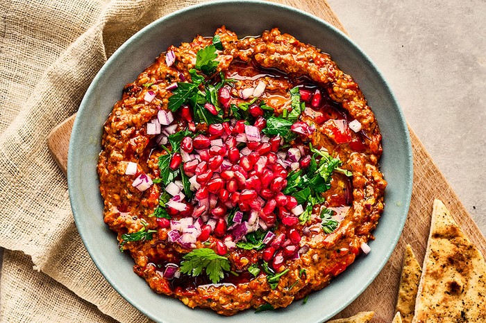 A bowl of muhammara topped with red onion, parsley and pomegranate seeds