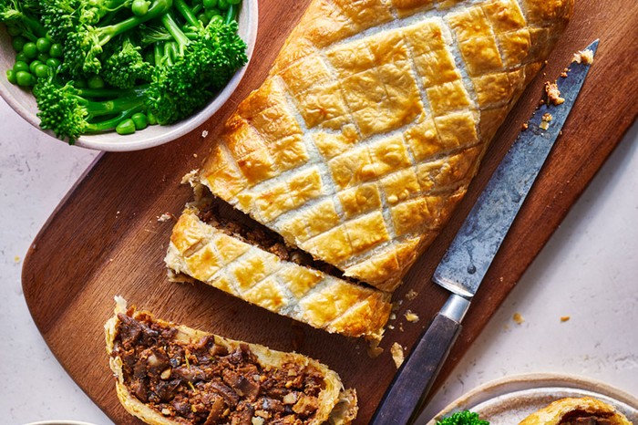 Mushroom wellington on a wooden board next to a bowl of broccoli and peas and another bowl of potatoes