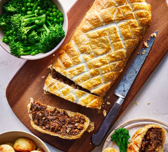 Mushroom wellington on a wooden board next to a bowl of broccoli and peas and another bowl of potatoes