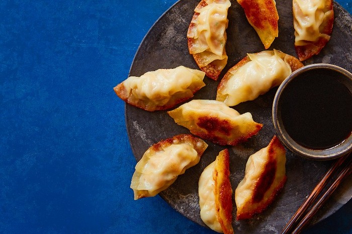 Nanami gyoza on a black plate with a pot of dipping sauce against a blue background