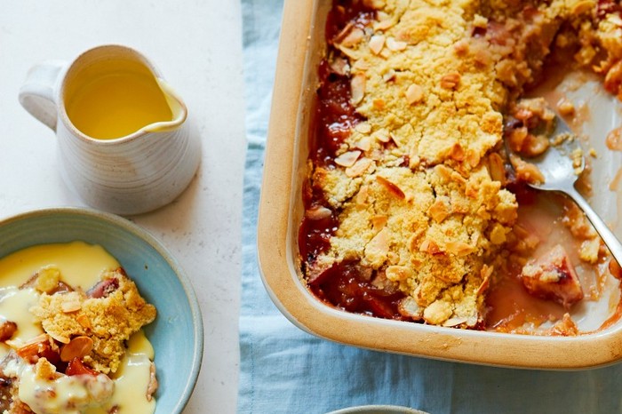 An oven dish of nectarine crumble on a blue cloth next to a small jug of custard