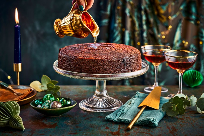 Gold jug pouring rum onto a CAribbean rum cake on a crystal cake stand