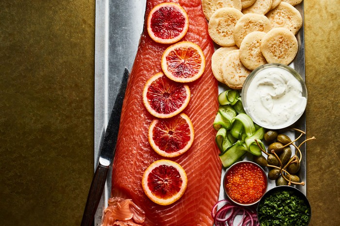 Large cured salmon fillet decorated with blood orange slices, served next to blinis, soured cream, cucumber, fish roe, olives, parsley and red onion