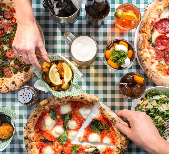 Two different hands reaching across the table for food, with three different Naples-style pizza, salad and drinks