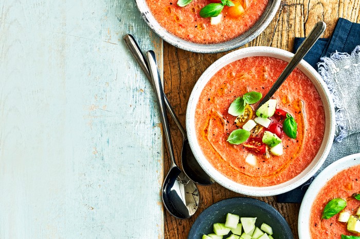 A blue wooden background topped with bowls of tomato soup, decorated with cubes of tomatoes, cucumber and basil leaves