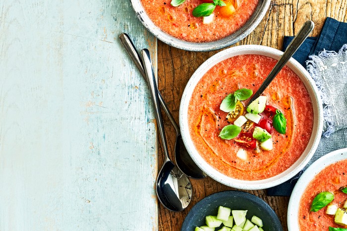 Vegetable and watermelon gazpacho A blue wooden background topped with bowls of tomato soup, decorated with cubes of tomatoes, cucumber and basil leaves