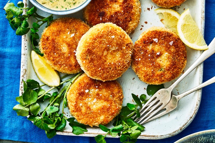 A white rectangular platter topped with golden fried crab cakes, with wedges of yellow lemon and green leaves