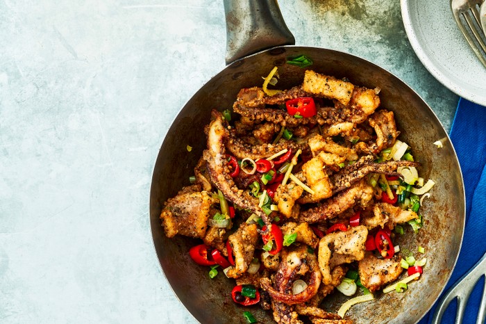 A frying pan filled with fried squid, red chillies and topped with green spring onions, set against a pale blue background