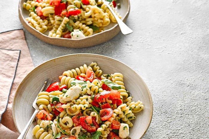 Two bowls filled with curly pasta, roast tomatoes, basil and white cheese balls