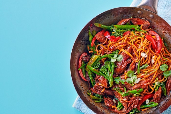 A frying pan filled dwith lamb, noodles, red pepper and green broccoli, on a blue background