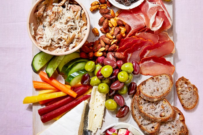 A serving board loaded with bread, cheese, pickles, cured meat and veg, with a knife on a pink background