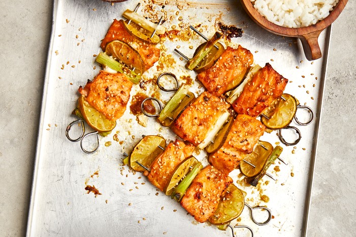 A metal tray topped with skewers pierced with chunks of salmon with spring onions and lime