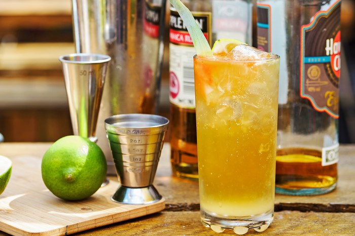 A wooden table topped with a rum cocktail, with silver cocktail shakers and a wedge of lime on the side