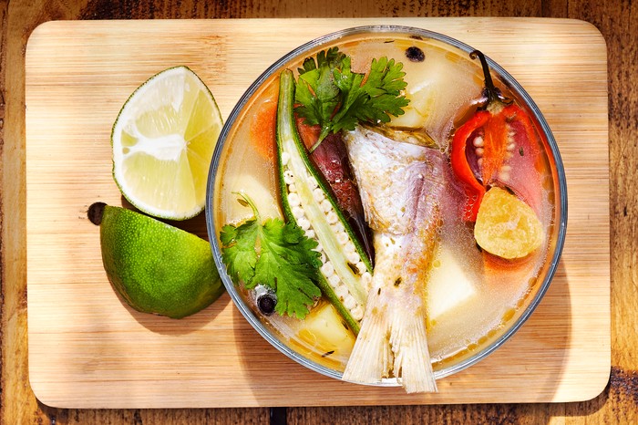 A wooden table topped with a bowl of fish soup with halves peppers, herbs and lime wedges