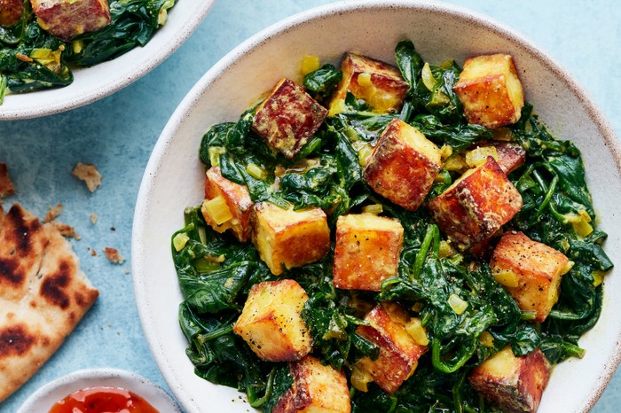 Two white bowls of golden cubes of paneer and wilted spinach on a blue background with a naan