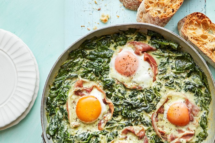 Metal Pan Filled with Creamy Spinach and Eggs Next to Slices of Ciabatta on a Blue Background