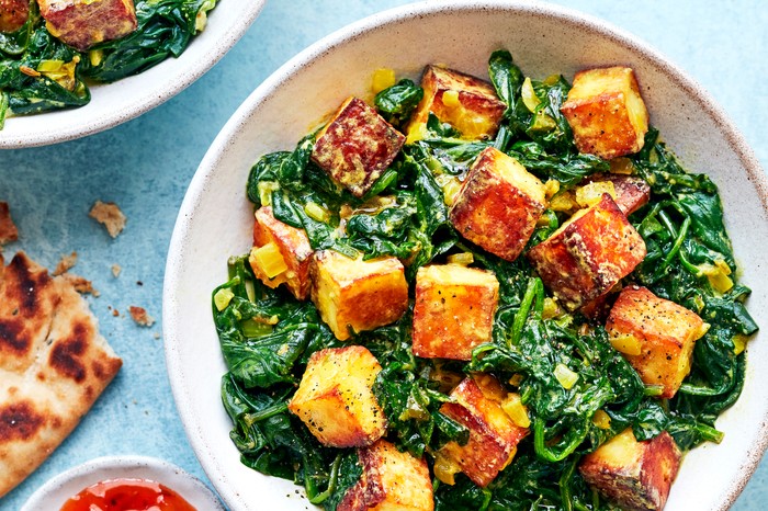Saag paneer Two white bowls of golden cubes of paneer and wilted spinach on a blue background with a naan