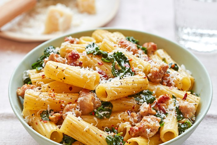 A bowl of rigatoni pasta with spinach and meat, with a plate of parmesan in the background on a white cloth