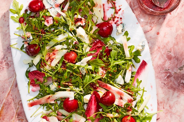 A white oval platter on pink mottled stone, topped with rocket leaves and cherries