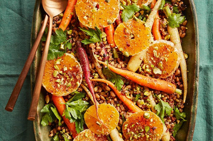 A green platter topped with grains, slices of orange, fermented carrots and crushed green pistachio nuts