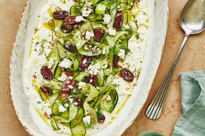 A white platter filled with ribboned cucumber, chopped black olives and crumbled feta on a stone background with a silver serving spoon