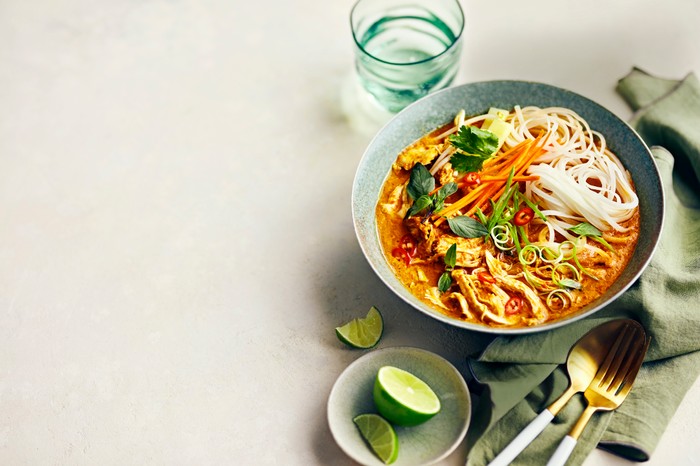 A bowl of chicken soup in a red-tinged broth with lime on the side