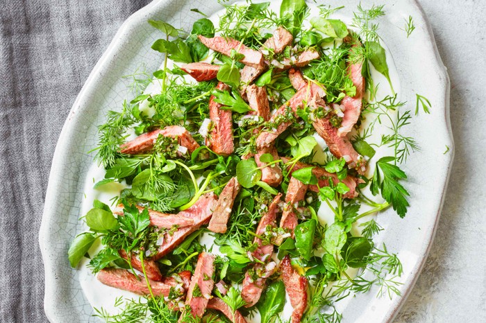 A white platter with green leaves and slices of pink lamb