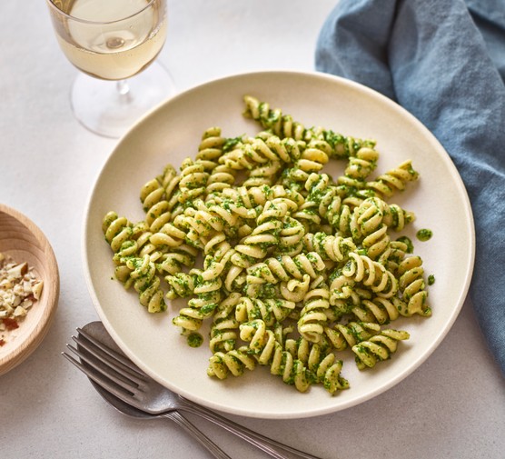 A bowl of fusilli pasta coated in green pesto next to a glass of wine and blue linen napkin